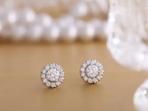 A pair of round diamond halo stud earrings on a gold-toned surface, with pearls and cut glass behind