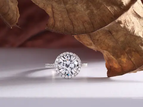 A round diamond halo ring on a white surface beside a dried leaf