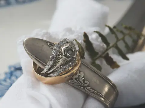 An old gold band and a vintage white gold diamond ring resting on an engraved silver spoon handle and white linen, with a fern leaf